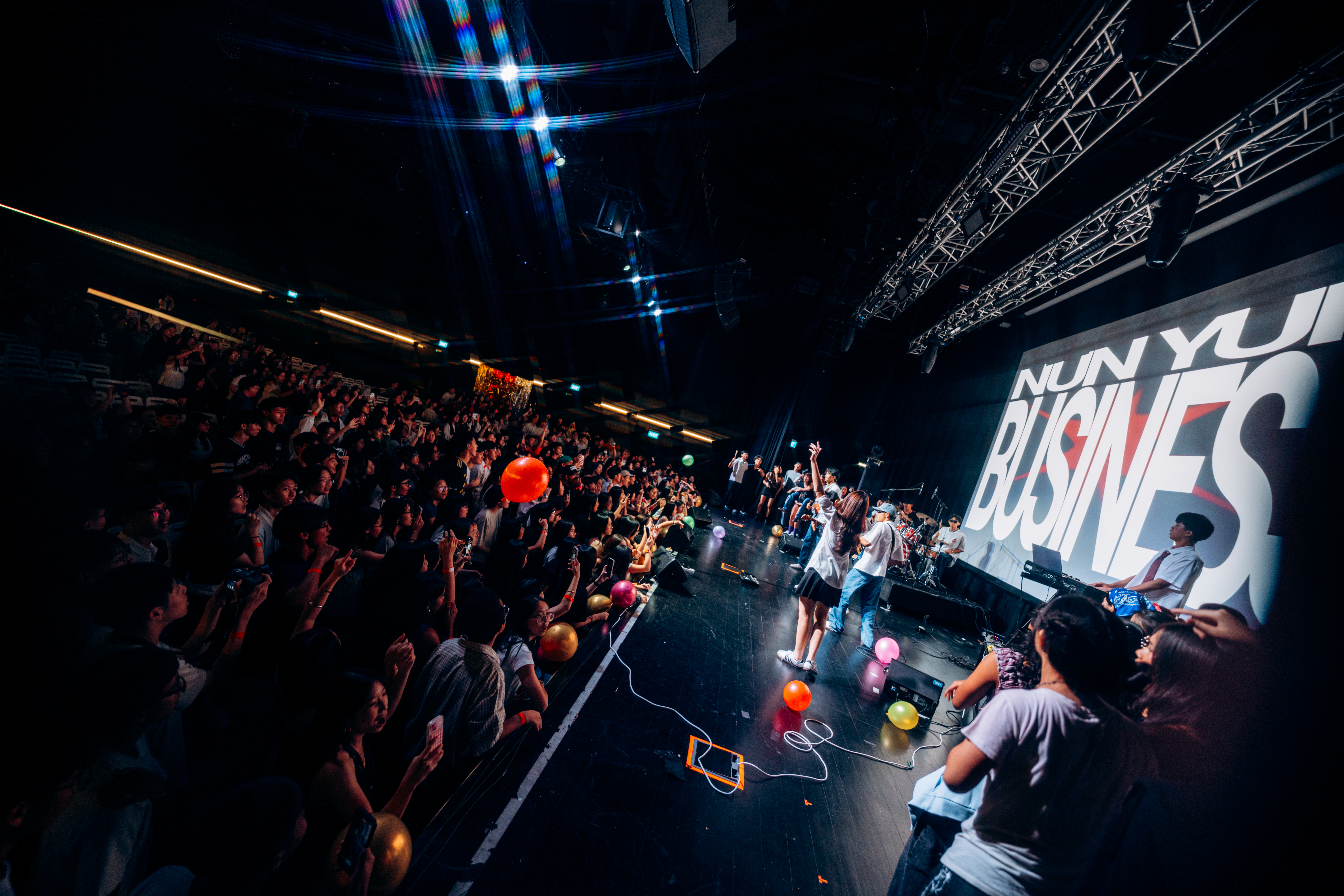 A packed crowd at an Amplify live show, band on stage under a Nun Yur Business backdrop